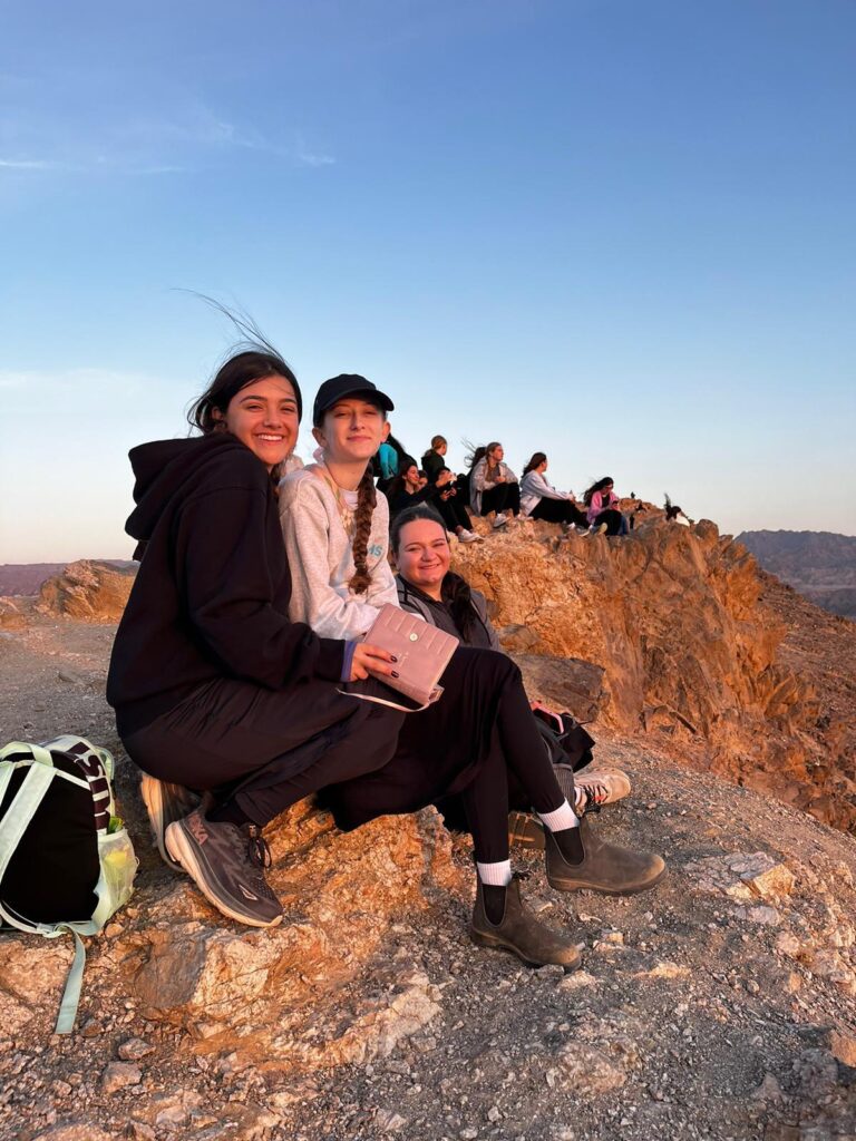Smiling friends sitting on rocky cliff at sunset