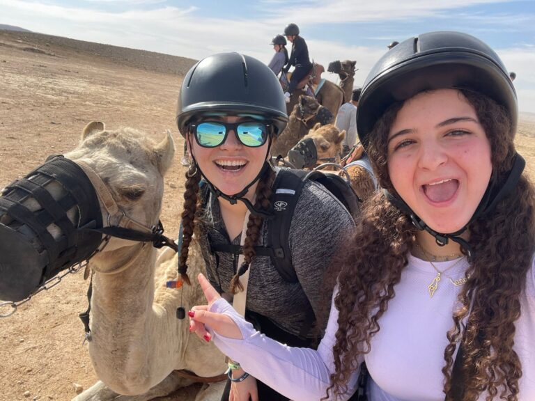 Two helmeted riders smiling beside camels